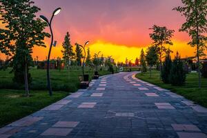 città parco nel presto estate o primavera con marciapiede, lanterne, giovane verde prato, alberi e drammatico nuvoloso cielo su un' tramonto o Alba. foto