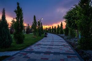 città parco nel presto estate o primavera con marciapiede, lanterne, giovane verde prato, alberi e drammatico nuvoloso cielo su un' tramonto o Alba. foto