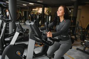 giovane donna con cuffie fare esercizi su Stazionario bicicletta nel un' Palestra o fitness centro. giovane sportivo donna nel Palestra ascolta musica a partire dal smartphone. donne fare cardio esercizi foto