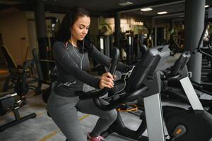 giovane donna con cuffie fare esercizi su Stazionario bicicletta nel un' Palestra o fitness centro. giovane sportivo donna nel Palestra ascolta musica a partire dal smartphone. donne fare cardio esercizi foto