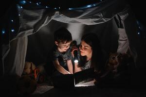 contento famiglia madre e bambino figlio lettura un' libro con un' torcia elettrica nel un' tenda a casa. famiglia concetto. foto