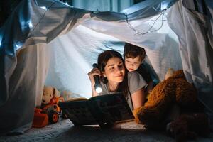 contento famiglia madre e bambino figlio lettura un' libro con un' torcia elettrica nel un' tenda a casa. famiglia concetto. foto