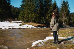 elegante ragazza in viaggio nel boschi. giovane donna nel cappello esplorando nel estate foresta, guardare a fiume. viaggio e voglia di girovagare concetto. spazio per testo. foto