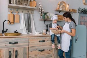 giovane contento mamma e sua bambino cucinare biscotti a casa nel il cucina. Natale fatti in casa Pan di zenzero. carino ragazzo con madre nel bianca uniforme e cappello cucinato cioccolato biscotti foto