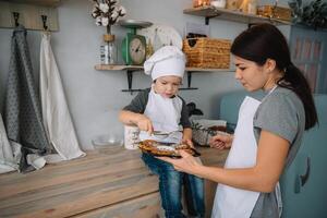 giovane contento mamma e sua bambino cucinare biscotti a casa nel il cucina. Natale fatti in casa Pan di zenzero. carino ragazzo con madre nel bianca uniforme e cappello cucinato cioccolato biscotti foto