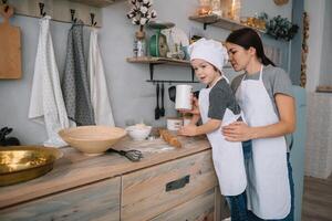 giovane contento mamma e sua bambino cucinare biscotti a casa nel il cucina. Natale fatti in casa Pan di zenzero. carino ragazzo con madre nel bianca uniforme e cappello cucinato cioccolato biscotti foto