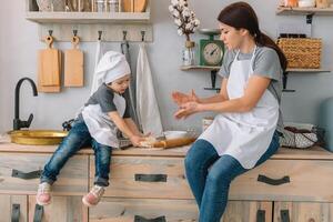 giovane contento mamma e sua bambino cucinare biscotti a casa nel il cucina. Natale fatti in casa Pan di zenzero. carino ragazzo con madre nel bianca uniforme e cappello cucinato cioccolato biscotti foto