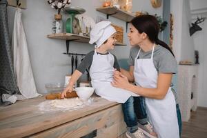 giovane contento mamma e sua bambino cucinare biscotti a casa nel il cucina. Natale fatti in casa Pan di zenzero. carino ragazzo con madre nel bianca uniforme e cappello cucinato cioccolato biscotti. foto