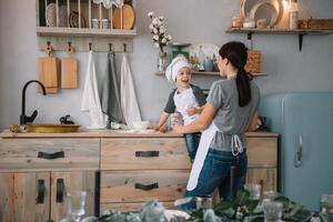 giovane contento mamma e sua bambino cucinare biscotti a casa nel il cucina. Natale fatti in casa Pan di zenzero. carino ragazzo con madre nel bianca uniforme e cappello cucinato cioccolato biscotti foto