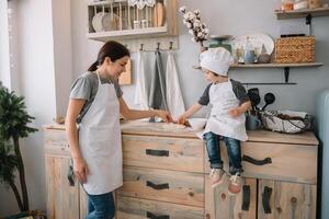 giovane contento mamma e sua bambino cucinare biscotti a casa nel il cucina. Natale fatti in casa Pan di zenzero. carino ragazzo con madre nel bianca uniforme e cappello cucinato cioccolato biscotti. foto