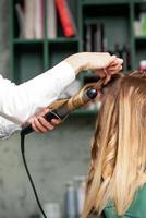 la creazione di riccioli con arricciatura ferri da stiro. parrucchiere fa un' acconciatura per un' giovane donna con lungo rosso capelli nel un' bellezza salone. foto