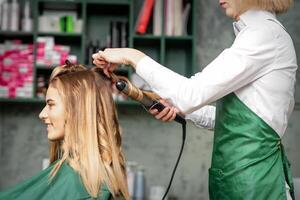 la creazione di riccioli con arricciatura ferri da stiro. parrucchiere fa un' acconciatura per un' giovane donna con lungo rosso capelli nel un' bellezza salone. foto