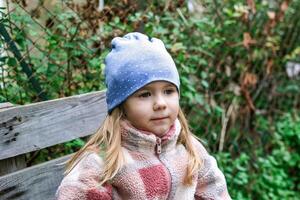 un' carino prescolastico ragazza con lungo biondo capelli e blu occhi seduta su un' di legno panchina nel il giardino indossare un' rosso e bianca vello giacca e blu berretto, guardare contento a il telecamera. foto