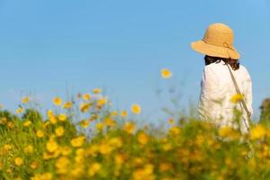 donna che indossa cappello di paglia che cammina nel campo di fiori gialli sotto il cielo blu in estate
