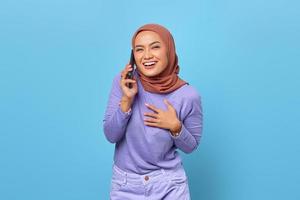 sorridente giovane donna asiatica che parla al telefono cellulare e mette le mani sul petto su sfondo blu foto
