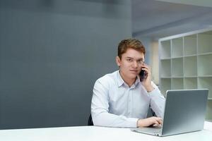 giovane professionale tipo conduce attività commerciale negoziati al di sopra di il Telefono mentre seduta a il suo all'avanguardia il computer portatile nel un' elegante ufficio foto