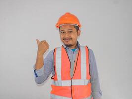 asiatico costruttore lavoratore uomo con sicurezza veste puntamento e mostrando con pollice su per il lato con contento viso sorridente foto