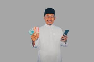 musulmano asiatico uomo sorridente contento mentre Tenere mobile Telefono e i soldi foto