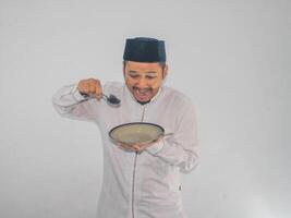 musulmano asiatico uomo guardare per vuoto mangiare piatto e Tenere cucchiaio con divertente espressione foto