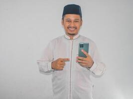 musulmano asiatico uomo sorridente e puntamento per mobile Telefono quello lui hold foto