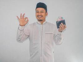 adulto musulmano asiatico uomo sorridente e dare ok dito cartello mentre Tenere i soldi foto