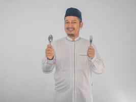 musulmano asiatico uomo sorridente contento con mano Tenere pranzo cucchiaio e forchetta foto