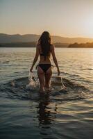 un' donna nel un' bikini in piedi nel il acqua a tramonto foto