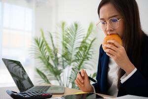 occupato e stanco donna d'affari mangiare pane e latte per pranzo a il scrivania ufficio e Lavorando per consegnare finanziario dichiarazioni per un' capo. oberati di lavoro e malsano per pronto pasti, bruciato concetto. foto