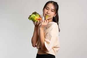 ritratto di un' contento giocoso asiatico ragazza mangiare fresco insalata a partire dal un' bicchiere ciotola dopo allenarsi a casa. giovane signora godendo salutare nutrizione e biologico cibo, avendo vegetariano pasto foto