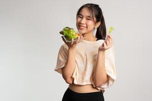 ritratto di un' contento giocoso asiatico ragazza mangiare fresco insalata a partire dal un' bicchiere ciotola dopo allenarsi a casa. giovane signora godendo salutare nutrizione e biologico cibo, avendo vegetariano pasto foto