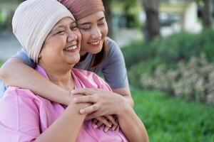 asiatico attento badante o infermiera e donna con cancro è seduta nel un' sedia a rotelle. nel il giardino. concetto di contento la pensione con cura a partire dal un' badante e risparmi e anziano Salute assicurazione. foto