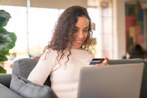 donna latina che utilizza laptop e mano che tiene la carta di credito per lo shopping sul divano foto