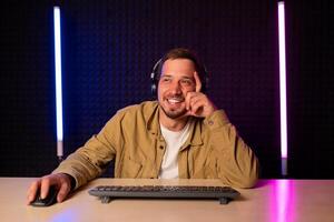 bello uomo nel camicia Abiti e cuffia seduta a un' tavolo nel un' camera con neon luci e giocando Giochi su il computer con un' Sorridi su il suo viso guardare a il schermo. foto