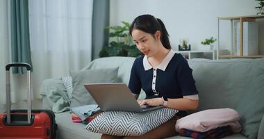 ritratto di asiatico adolescente donna seduta su divano utilizzando il computer portatile per preparare prenotazione Hotel e aereo biglietto per viaggio. zaino in spalla viaggio concetto. foto