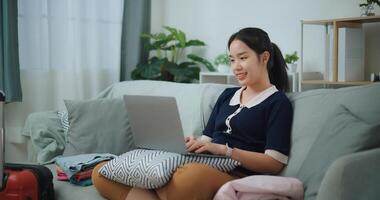 lato Visualizza di asiatico adolescente donna seduta su divano utilizzando il computer portatile per preparare prenotazione Hotel e aereo biglietto per viaggio. zaino in spalla viaggio concetto. foto