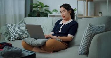 ritratto di asiatico adolescente donna seduta su divano utilizzando il computer portatile per preparare prenotazione Hotel e aereo biglietto per viaggio. zaino in spalla viaggio concetto. foto