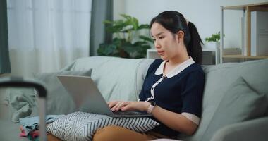 lato Visualizza di asiatico adolescente donna seduta su divano utilizzando il computer portatile per preparare prenotazione Hotel e aereo biglietto per viaggio. zaino in spalla viaggio concetto. foto