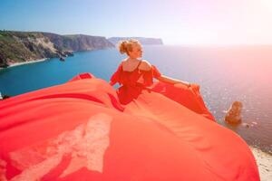 donna mare rosso vestire. bionda con lungo capelli su un' soleggiato riva del mare nel un' rosso fluente vestire, indietro Visualizza, seta tessuto agitando nel il vento. contro il fondale di il blu cielo e montagne su il spiaggia. foto