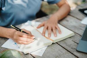 avvicinamento Visualizza di un' persona scrittura nel un' taccuino con un' penna, con un' il computer portatile per il lato su un' di legno all'aperto tavolo. foto