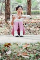 donna seduto su un' parco sentiero sembra a sua fitness inseguitore mentre Tenere un' smartphone, con sparpagliato autunno le foglie nel il sfondo. foto