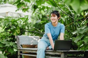 contento donna seduta su un' di legno panca, Lavorando su un' il computer portatile nel un' vivace giardino, godendo a distanza opera. foto