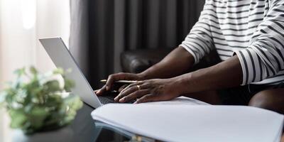 africano americano uomo Lavorando con il computer portatile computer chiamata e conferenza, a distanza opera mentre seduta a divano nel vivente camera. nero tipo fare libero professionista opera a casa ufficio foto