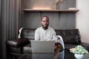 africano americano uomo Lavorando con il computer portatile computer a distanza mentre seduta a divano nel vivente camera. nero tipo fare libero professionista opera a casa ufficio foto