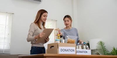 donazione e Due donna volontario asiatico di contento Imballaggio cibo nel scatola a casa. beneficenza foto