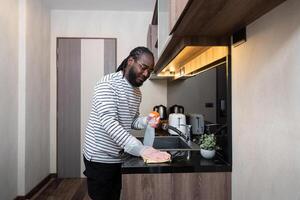 africano americano uomo nel gomma da cancellare guanti pulire polvere a partire dal tavolo nel cucina, lavori di casa, pulizia, stile di vita, domestico foto