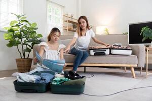 giovane asiatico donna Imballaggio Abiti per il valigia seduta su il divano. preparazione per il estate vacanza. Due donne siamo pianificazione un' viaggio e porzione per preparare bagaglio per viaggio foto