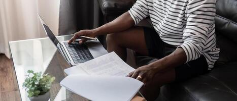 africano americano uomo Lavorando con il computer portatile computer a distanza mentre seduta a divano nel vivente camera. nero tipo fare libero professionista opera a casa ufficio foto