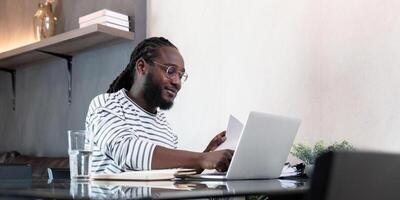 africano americano uomo Lavorando con il computer portatile computer a distanza mentre seduta a bicchiere tavolo nel vivente camera. nero tipo fare libero professionista opera a casa ufficio foto