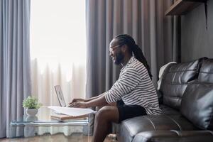 africano americano uomo Lavorando con il computer portatile computer a distanza mentre seduta a divano nel vivente camera. nero tipo fare libero professionista opera a casa ufficio foto