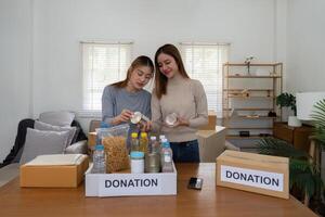 donazione e Due donna volontario asiatico di contento Imballaggio cibo nel scatola a casa. beneficenza foto